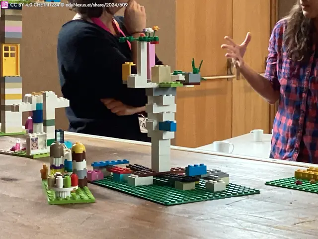 Zwei Personen diskutieren über LEGO-Kreationen auf einem Tisch. Links steht eine bunte Struktur mit Türmen, rechts eine hohe Säule mit bunten Elementen und Pflanzen oben.