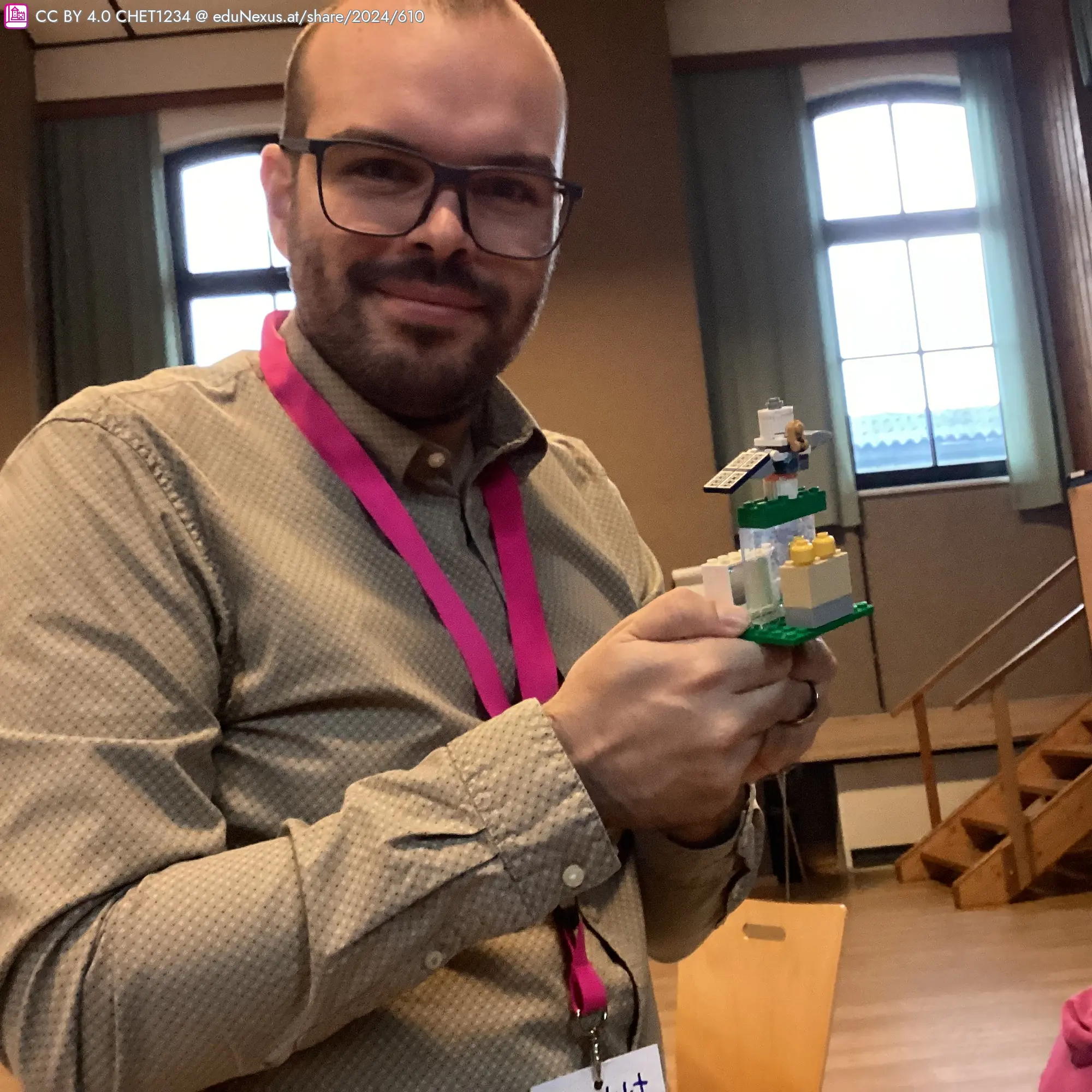 Mann mit Brille hält ein grünes Lego-Modell in der Hand, das aus mehreren Ebenen besteht. Er trägt ein kariertes Hemd und ein pinkes Namensband.