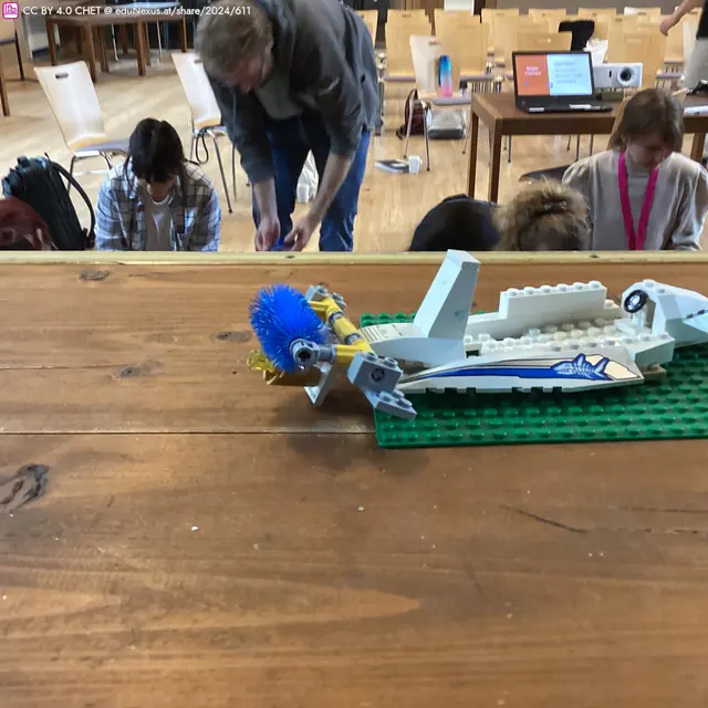 Ein LEGO-Modell eines Flugzeugs mit blauer und gelber Verzierung auf einem grünen Grund. Im Hintergrund sind mehrere Personen und Tische in einem Raum zu sehen.