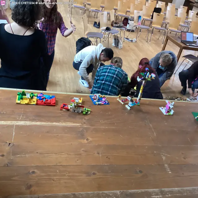 Menschen, die auf dem Boden sitzen und mit Lego-Steinen spielen, während einige Lego-Kreationen auf einem Tisch sichtbar sind.