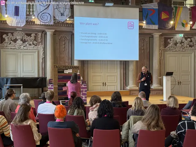 Konferenzraum mit Publikum, einer Person, die spricht, und einer Präsentationsfolie. Bunte Banner hängen an der Wand.