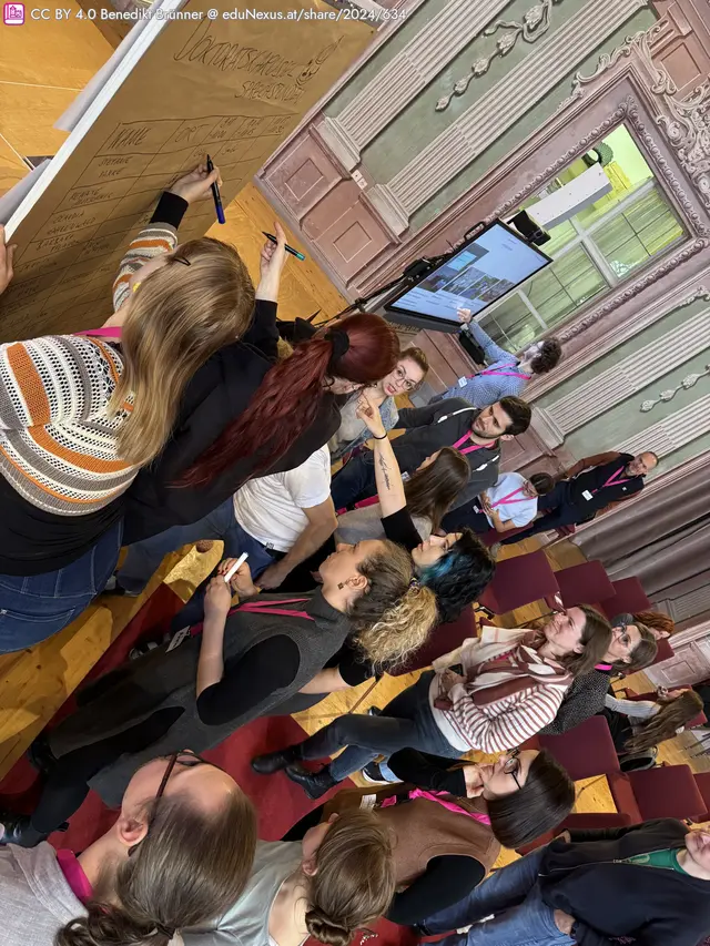 Gruppendiskussion in einem Saal, Teilnehmer schreiben an einem großen Blatt Papier, einige nutzen Smartphones, pinke Namensschilder sichtbar, historische Architektur im Hintergrund.