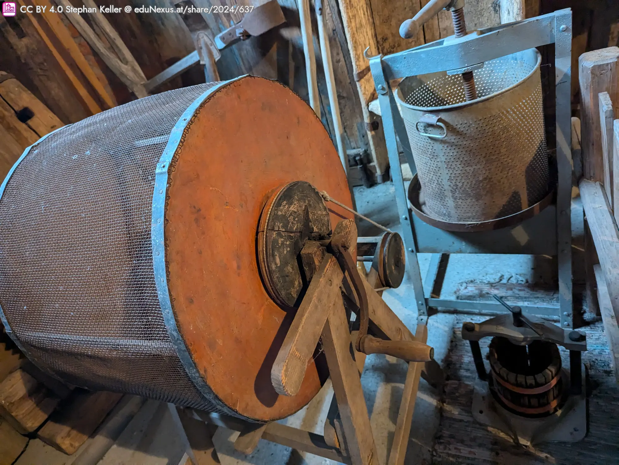 Eine alte Holzmaschine mit einem großen, runden Gitterbehälter und einem Handrad, daneben ein kleinerer, metallener Behälter auf einem Gestell.