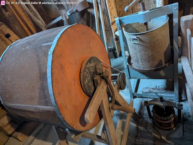 Eine alte Holzmaschine mit einem großen, runden Gitterbehälter und einem Handrad, daneben ein kleinerer, metallener Behälter auf einem Gestell.