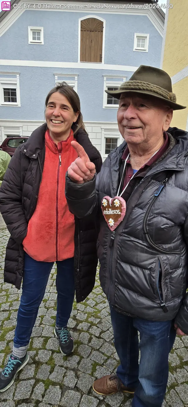 Zwei Personen stehen auf einem gepflasterten Platz vor einem blauen Gebäude. Die Frau trägt einen roten Pullover und eine dunkle Jacke, der Mann hat einen Hut auf und hält ein herzförmiges Lebkuchenherz.