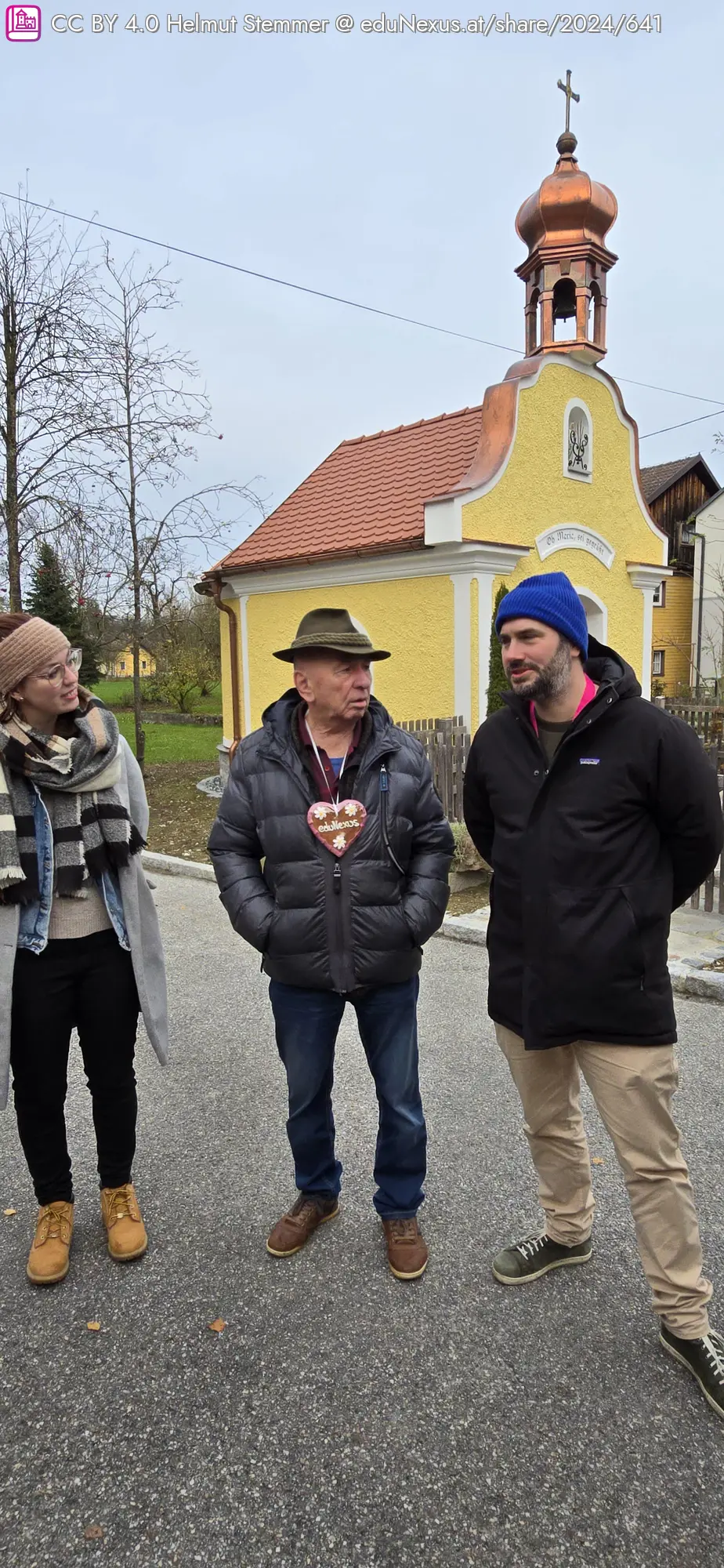 Drei Personen stehen auf einer Straße vor einer gelben Kirche mit einer Kupferkuppel. Die Frau links trägt einen Schal und eine gestreifte Jacke. Der Mann in der Mitte trägt einen Hut und einen dicken Mantel mit einer Herz-Anstecknadel. Der Mann rechts hat einen blauen Hut und eine schwarze Jacke.