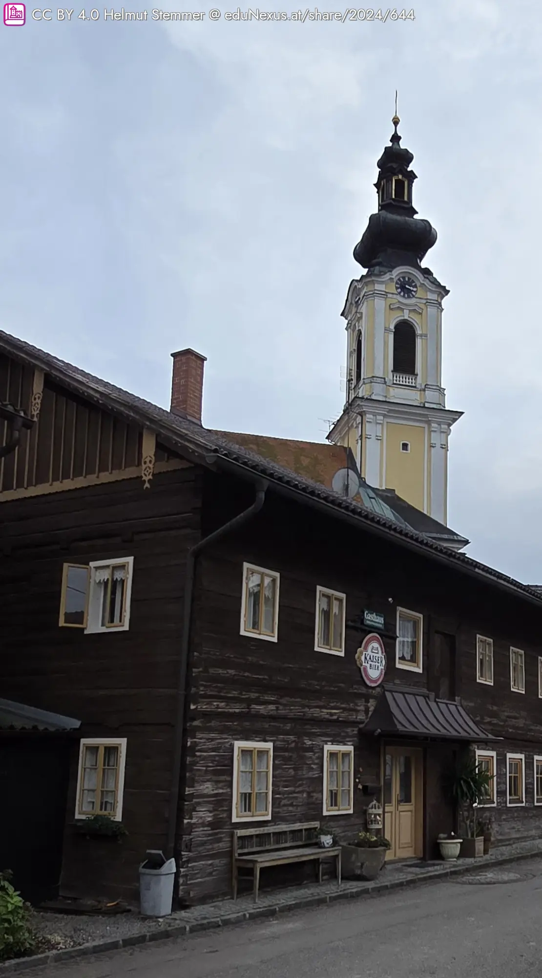 Holzgebäude mit mehreren Fenstern und einem Schild „Kaiser Bier“ im Vordergrund; im Hintergrund ein Kirchturm mit einer goldenen Kuppel und Uhr.