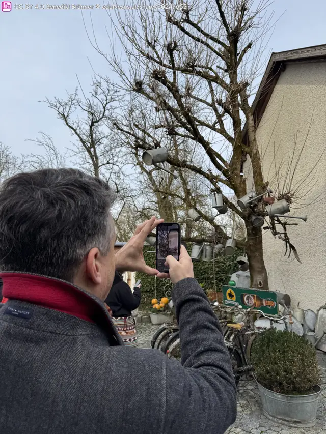 Mann fotografiert mit Smartphone einen Baum, an dem Gießkanne hängen. Im Hintergrund sind Fahrräder und Pflanzen zu sehen.