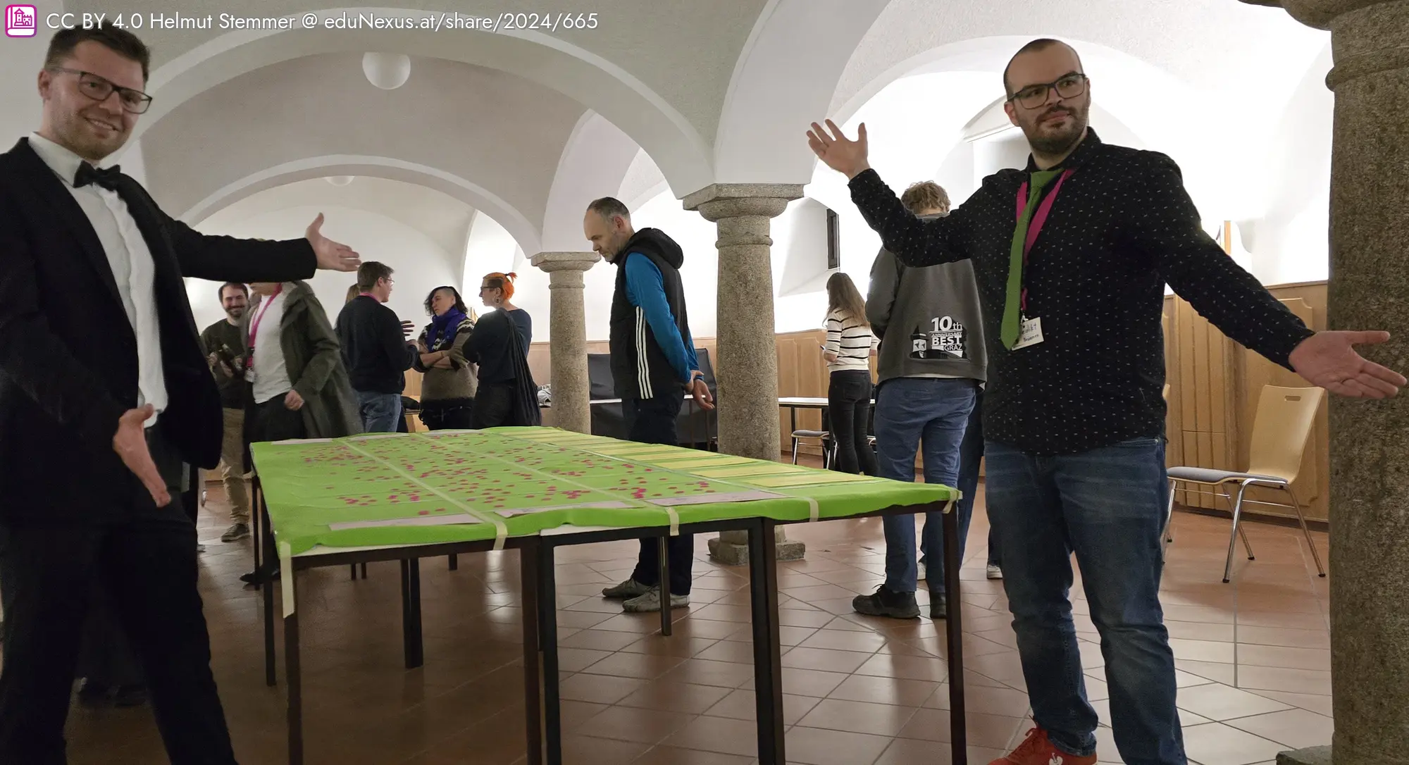 Zwei Männer in formeller Kleidung stehen an einem Tisch mit grünem Tischdecken. Im Hintergrund sind mehrere Personen und Säulen sichtbar.