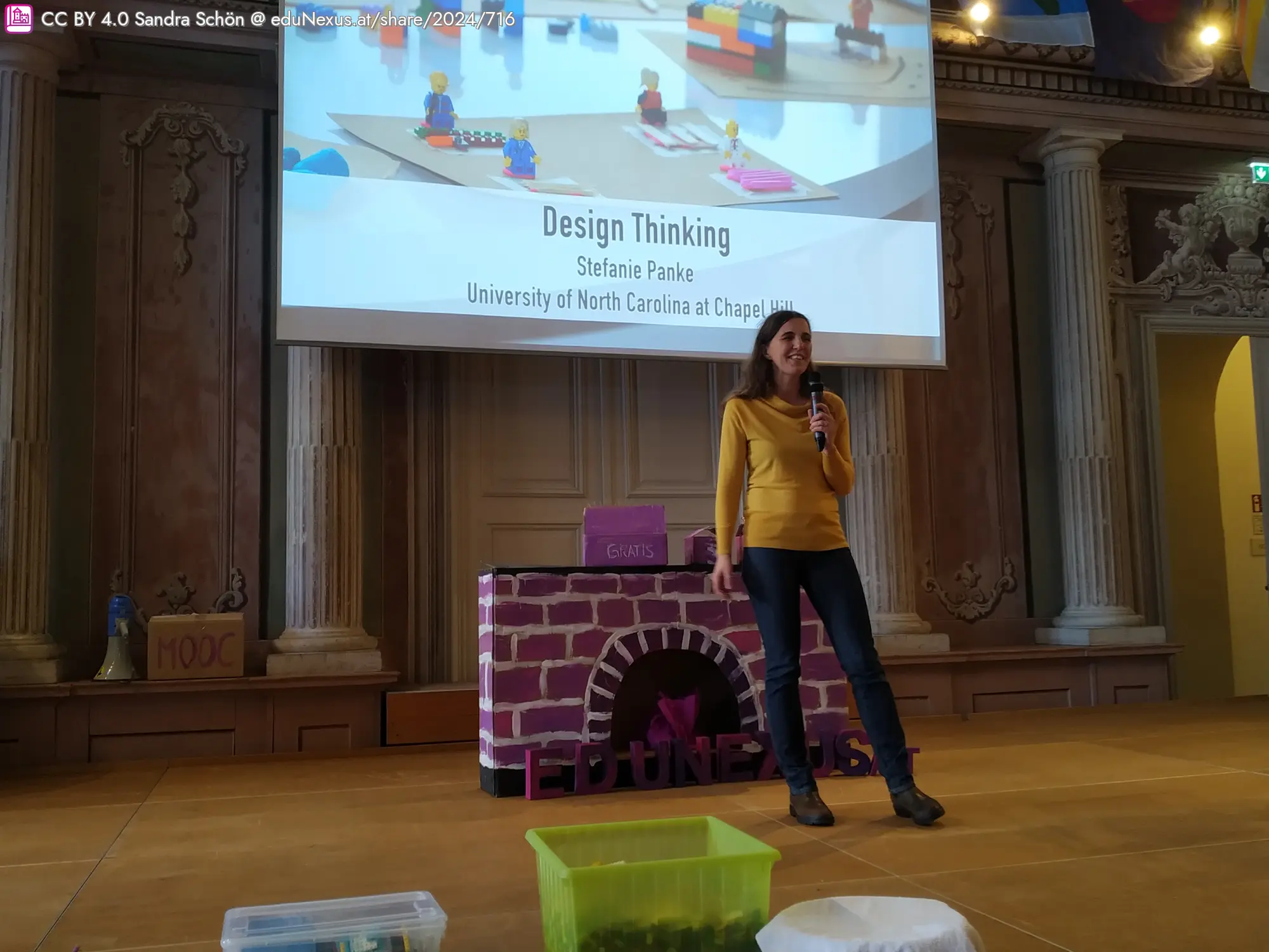 Frau mit dunklen Haaren und gelbem Oberteil steht auf einer Bühne vor einer Präsentation über Design Thinking. Im Hintergrund sind Lego-Figuren und ein Tisch mit bunten Behältern sichtbar.