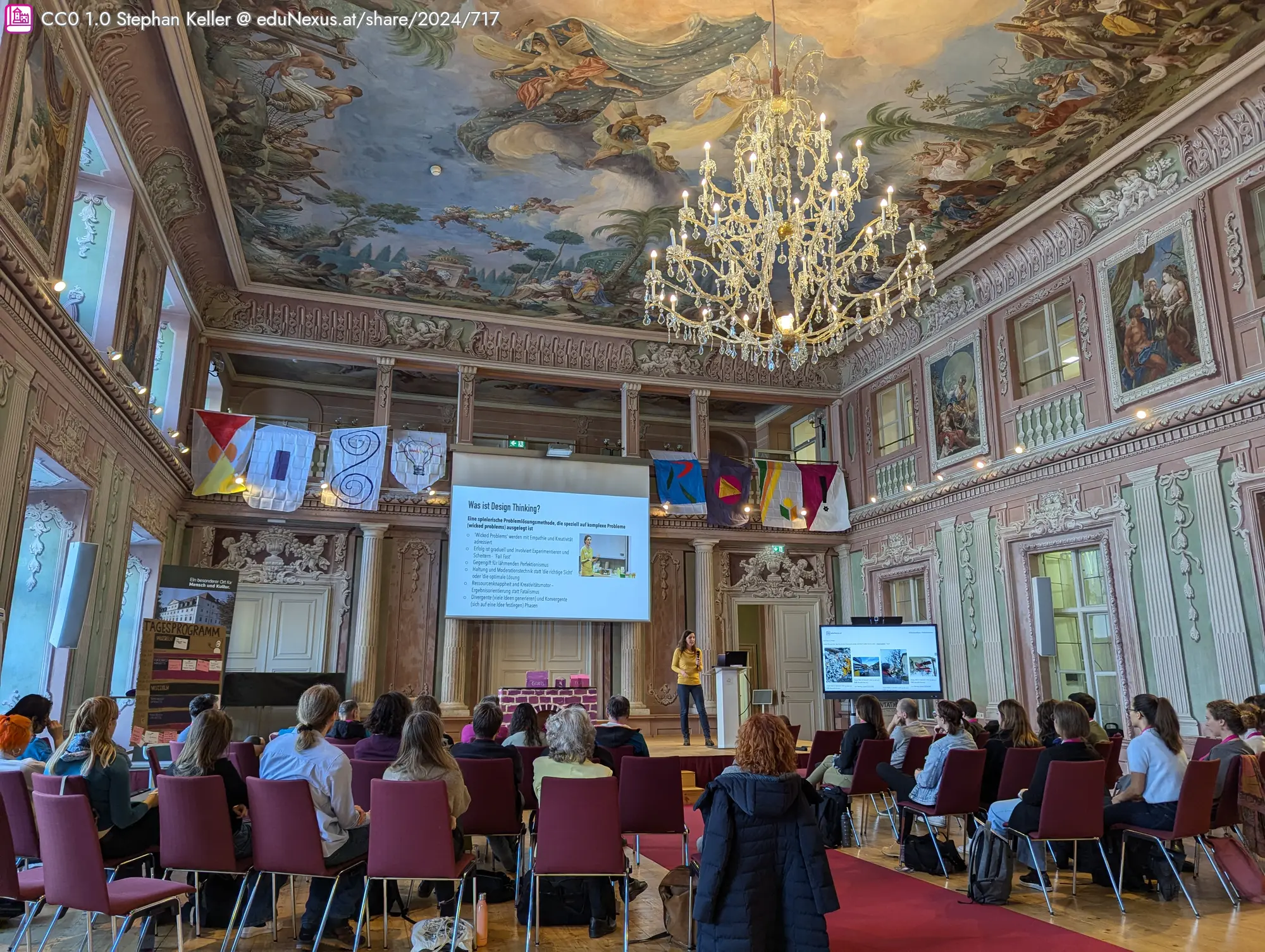 Ein historischer Raum mit einem kunstvoll bemalten Decke, einem großen Kronleuchter und bunten Fahnen. Eine Person steht am Podium vor einer Präsentation, während das Publikum in roten Stühlen sitzt.