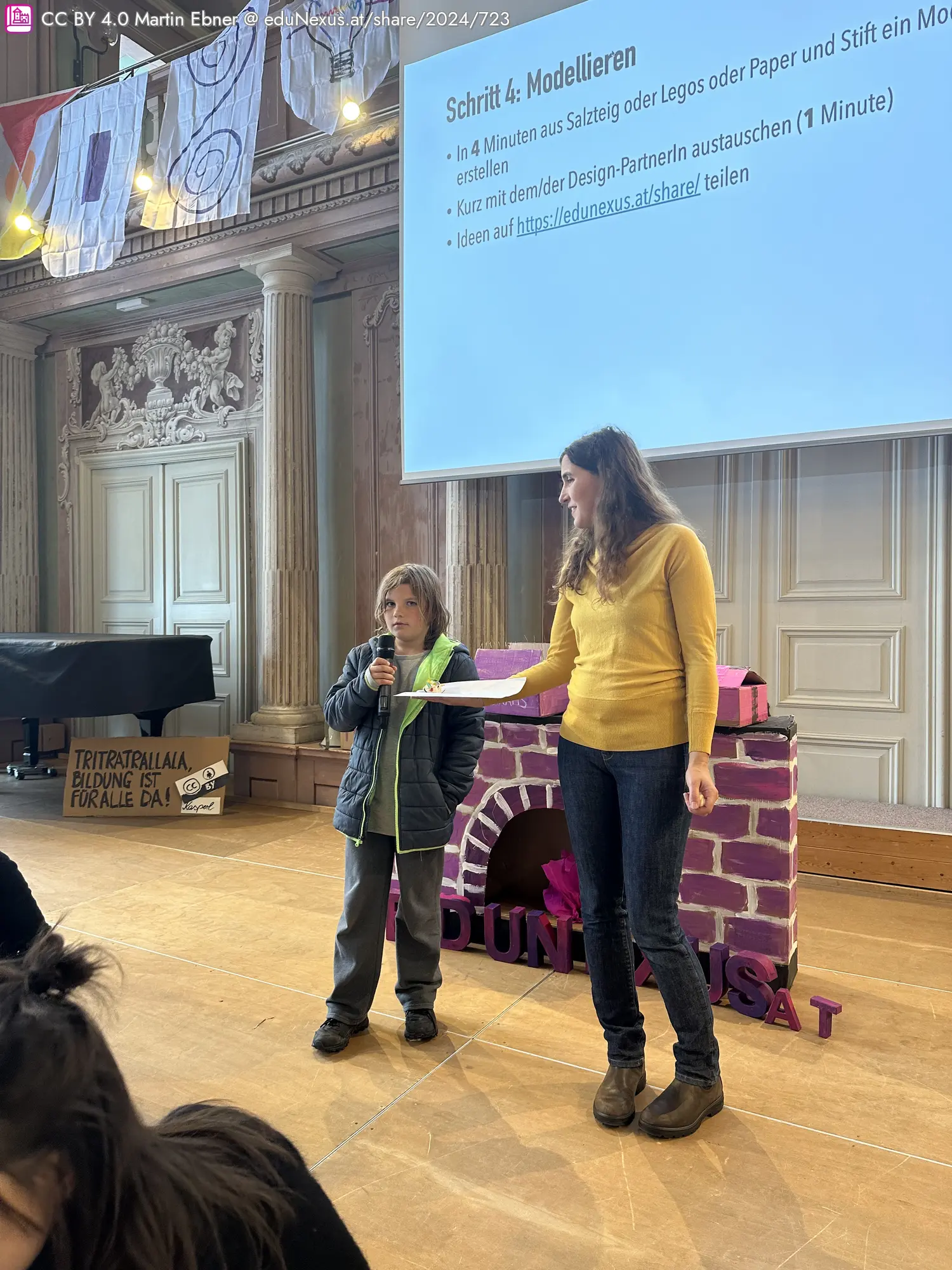 Ein Junge mit Mikrofon steht neben einer Frau in gelbem Pullover, die ein Blatt Papier hält. Im Hintergrund ist eine Präsentation an der Wand und eine pinkfarbene Struktur sichtbar.