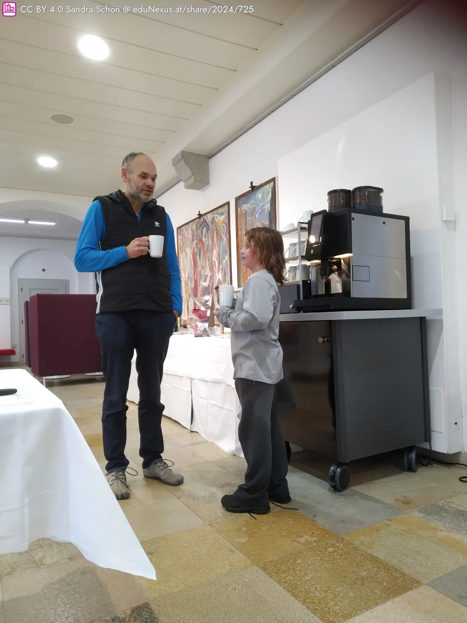 Ein Mann und ein Kind stehen in einem Raum mit einem Kaffeeautomaten. Beide halten Tassen. Im Hintergrund sind mehrere Kunstwerke an der Wand.