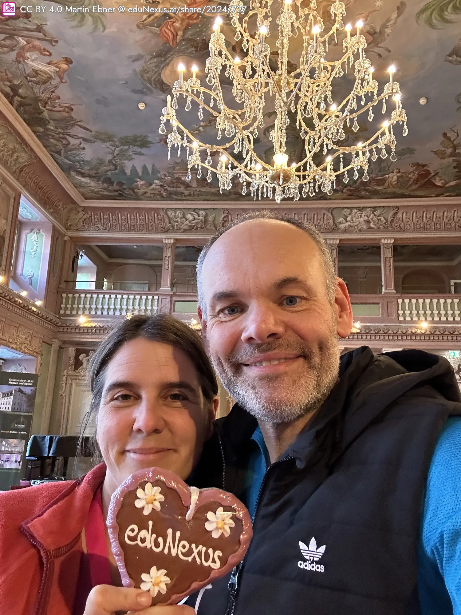 Zwei Personen posieren für ein Selfie in einem opulenten Raum mit einem Kristalllüster im Hintergrund. Eine Person hält ein herzförmiges Lebkuchenherz mit der Aufschrift „eduNexus“.