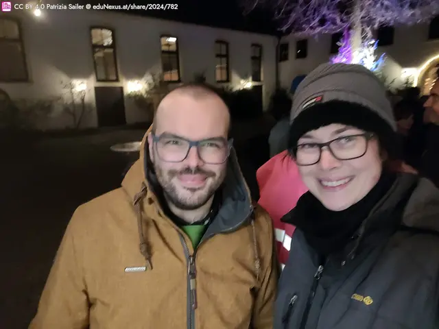 Zwei Personen stehen lächelnd vor einem beleuchteten Gebäude in der Nacht. Die Person links trägt eine braune Jacke, die Person rechts eine schwarze Mütze und einen dunklen Schal.