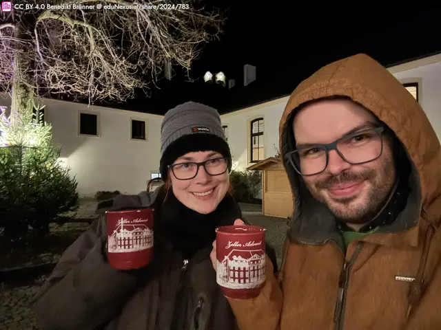 Zwei Personen im Freien halten Tassen in der Hand. Eine Person trägt eine graue Mütze und eine schwarze Jacke, die andere trägt eine braune Kapuzenjacke. Im Hintergrund sind Bäume und ein beleuchtetes Gebäude zu sehen.