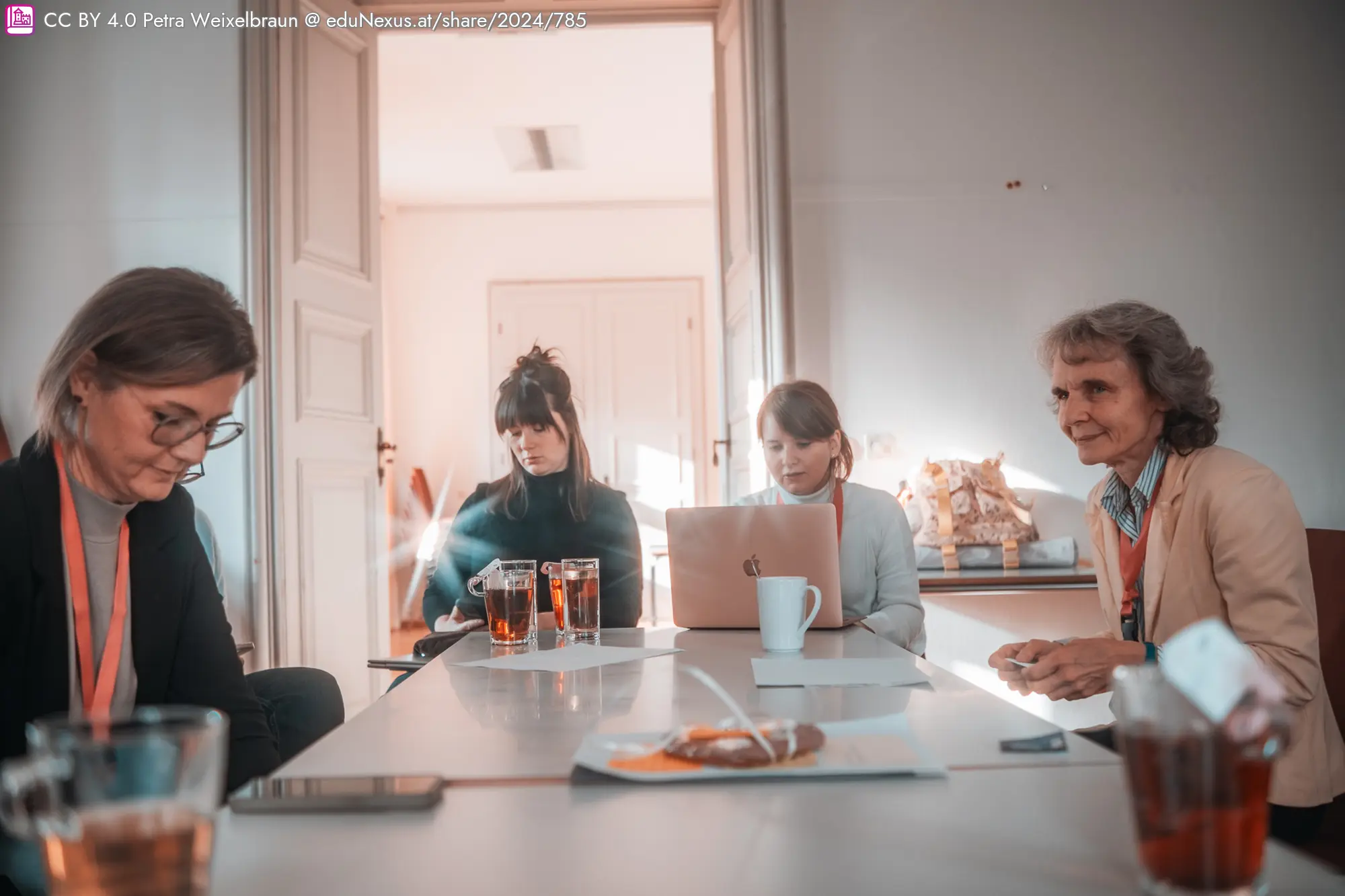 Vier Frauen sitzen an einem Tisch, zwei mit Getränken, eine arbeitet am Laptop. Heller Raum mit einer Tür im Hintergrund.