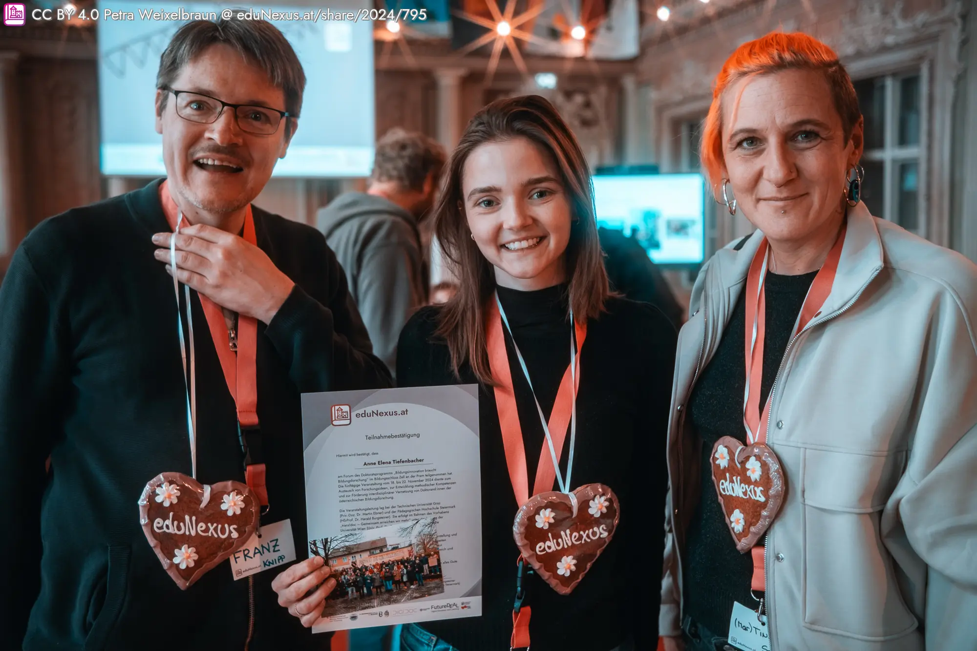 Drei Personen lächeln und stehen zusammen. Sie tragen pinke Lanyards mit Namensschildern und halten herzförmige Lebkuchen mit der Aufschrift „eduNexus“. Eine Person hält ein Zertifikat in der Hand. Hintergrund mit verschwommenen Personen und einem Bildschirm.