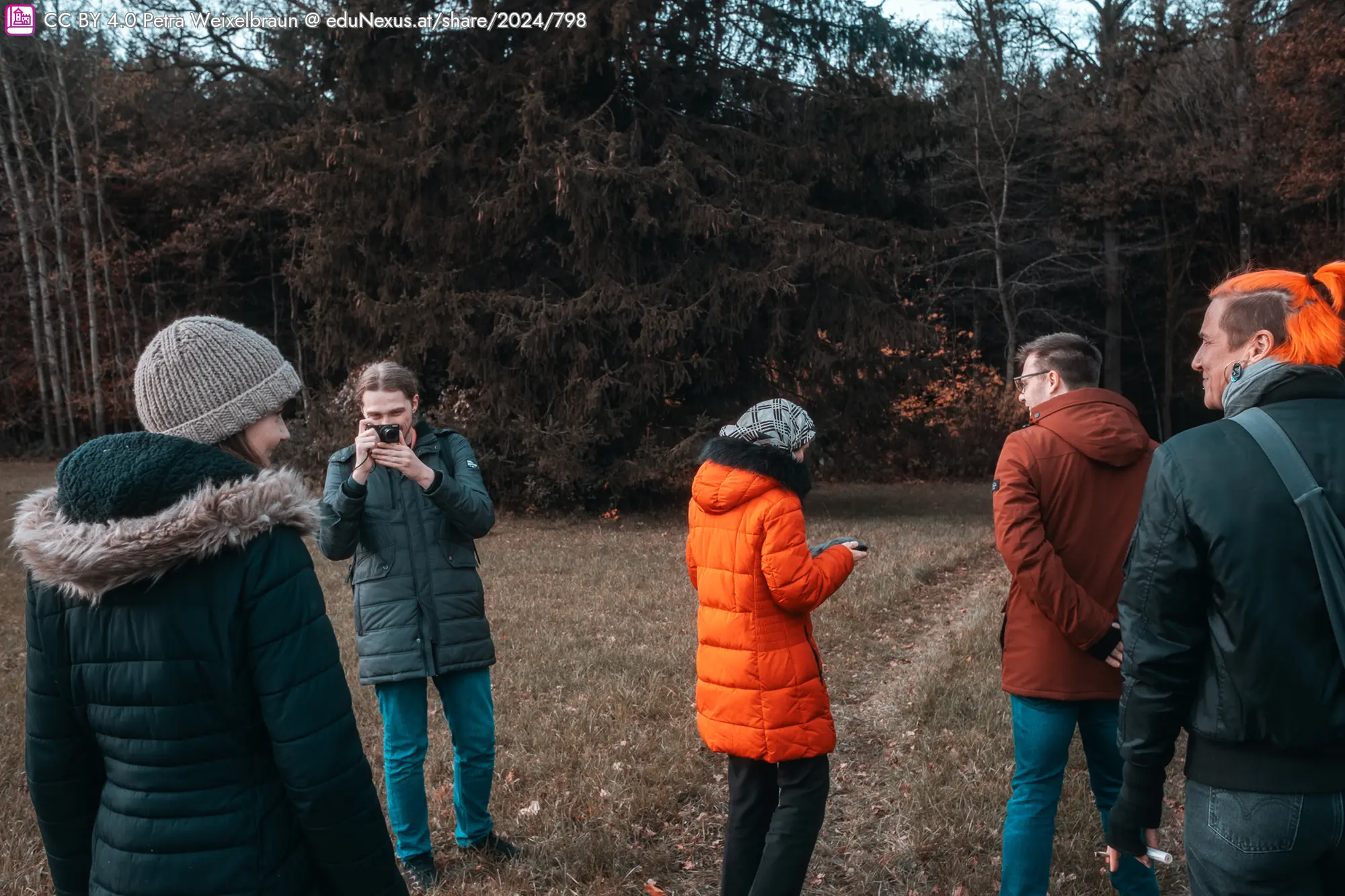 Sechs Personen in Winterjacken stehen auf einem Feld, umgeben von Bäumen. Eine Person in einem orangefarbenen Mantel hält ein Smartphone.