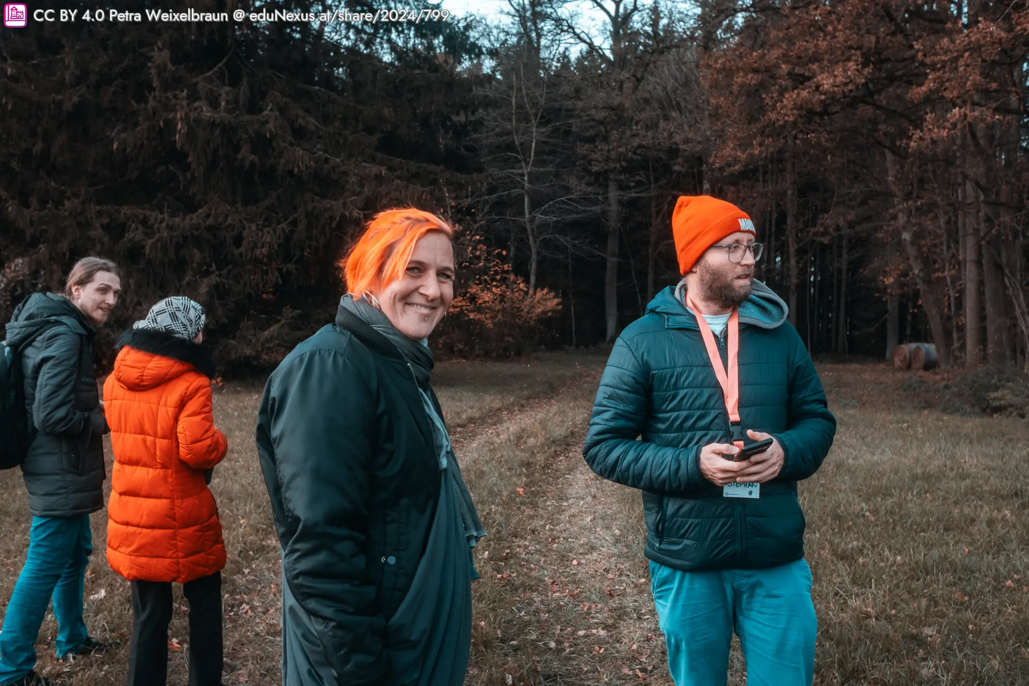 Vier Personen stehen auf einem Waldweg. Eine Person mit orangefarbenem Haar und einem schwarzen Mantel lächelt. Eine andere Person trägt eine orange Mütze und hält ein Smartphone. Die Umgebung zeigt Bäume und herbstliche Farben.