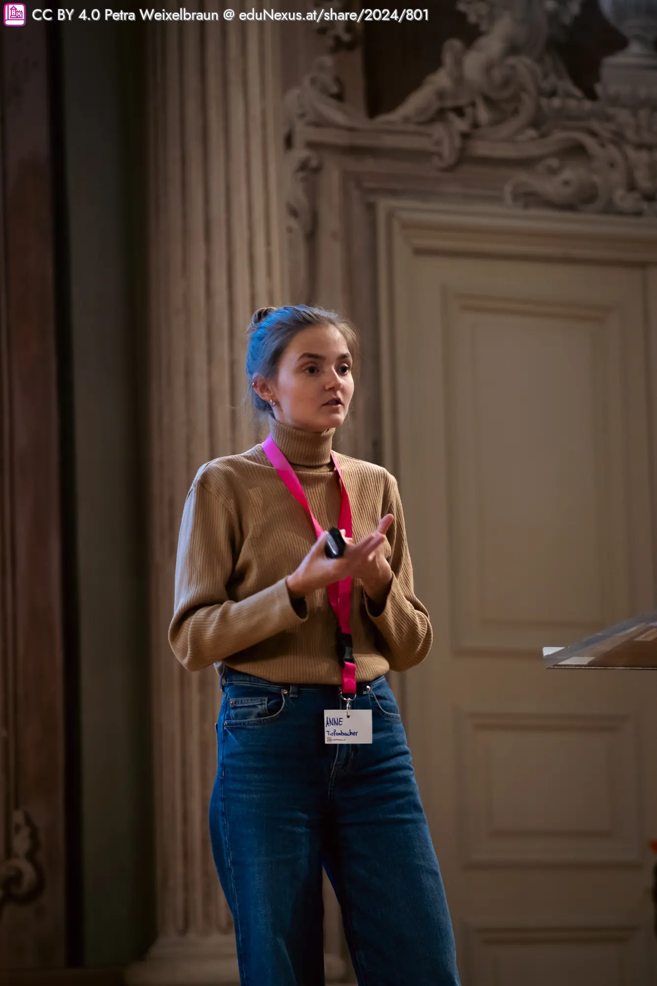 Eine junge Frau in einem beigen Oberteil und blauen Jeans steht vor einem Podium. Sie hat einen pinken Ausweis um den Hals und gestikuliert mit den Händen. Im Hintergrund sind dekorative Wände und eine Tür zu sehen.