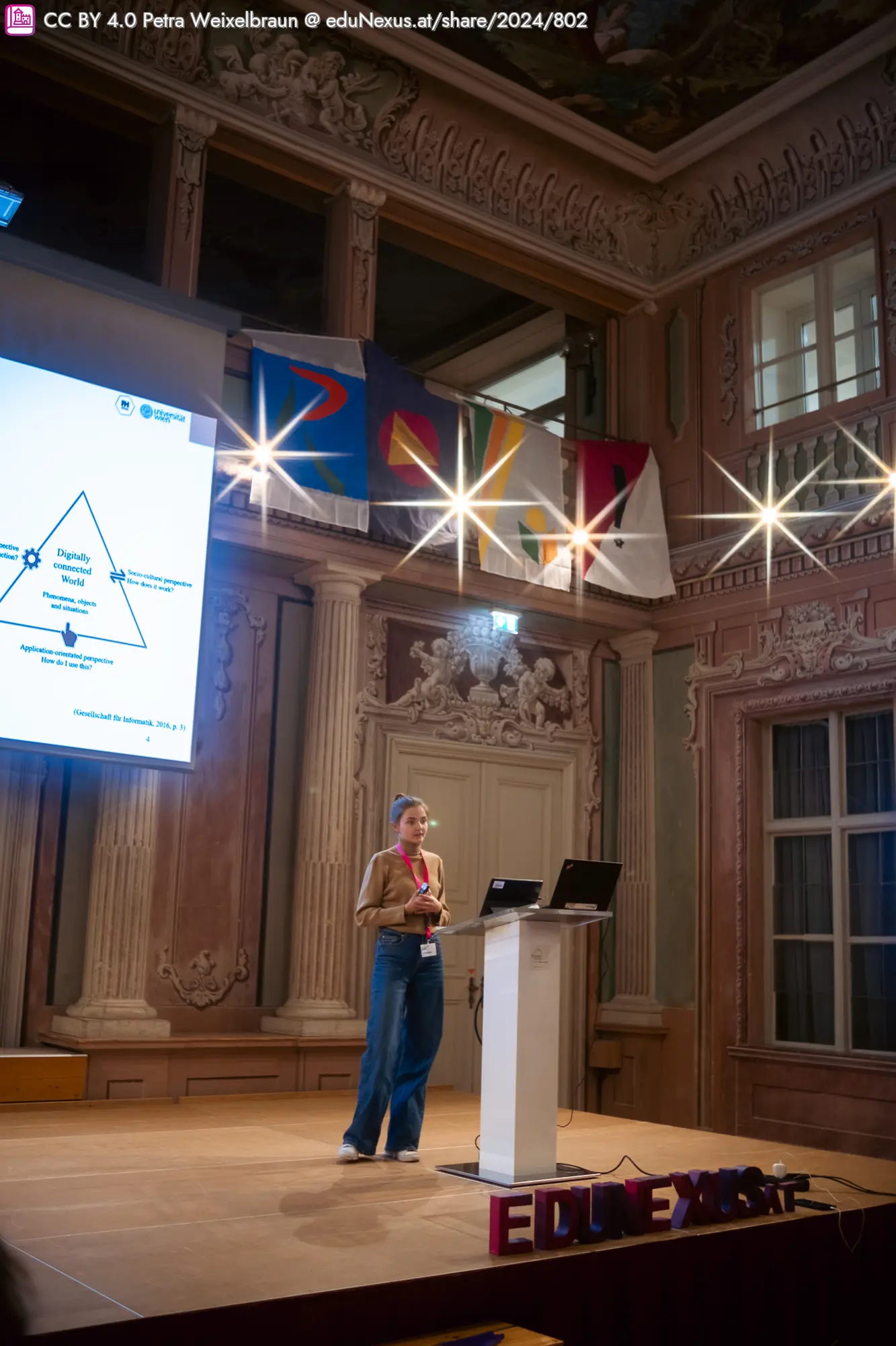 Frau mit blonden Haaren steht an einem Podium, präsentiert vor einem Bildschirm mit Diagramm. Im Hintergrund hängen mehrere Flaggen.