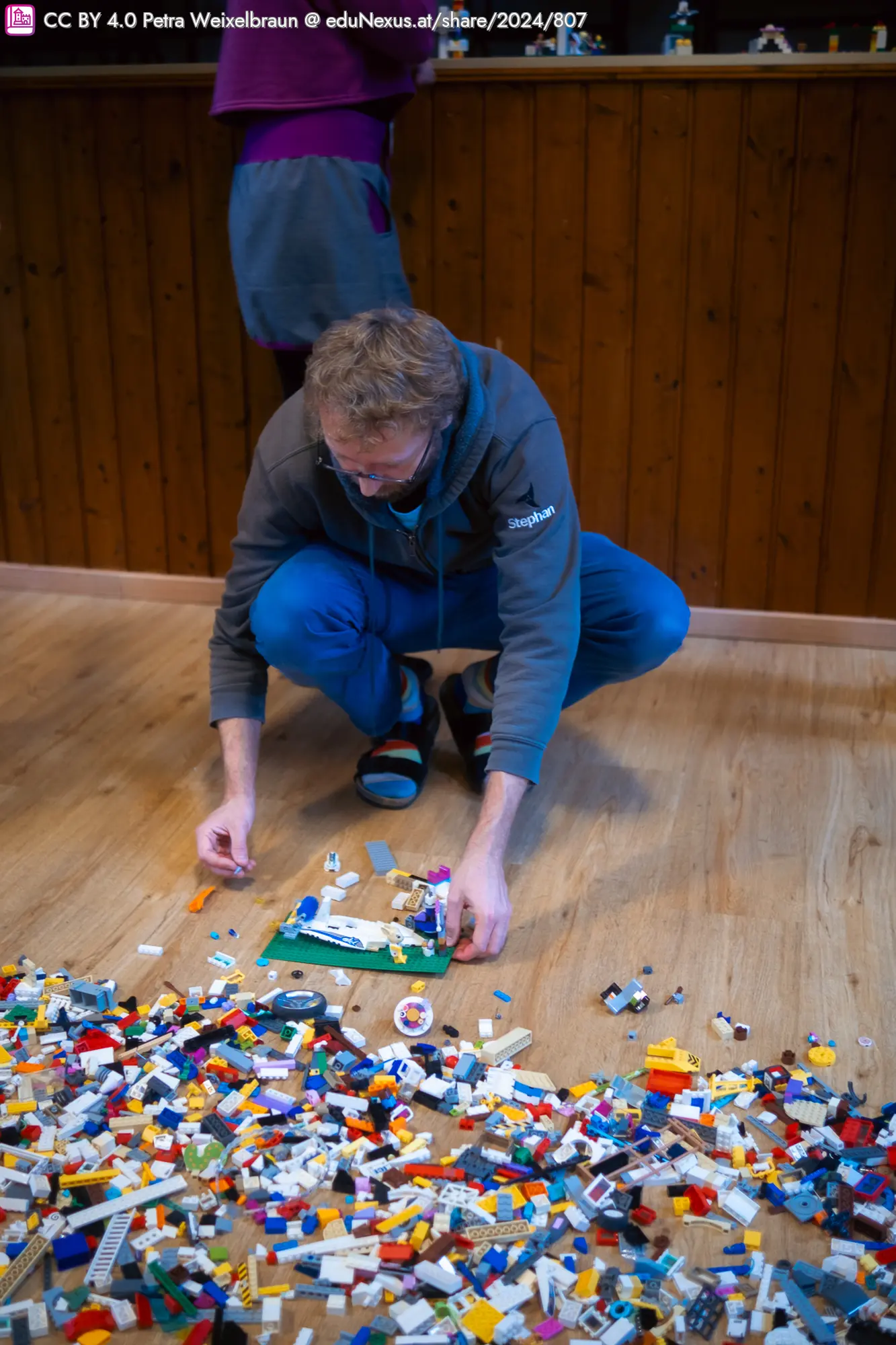 Mann in blauer Hose und grauem Hoodie, der auf dem Boden sitzt und mit bunten Lego-Steinen spielt. Um ihn herum liegt ein großer Haufen Lego.