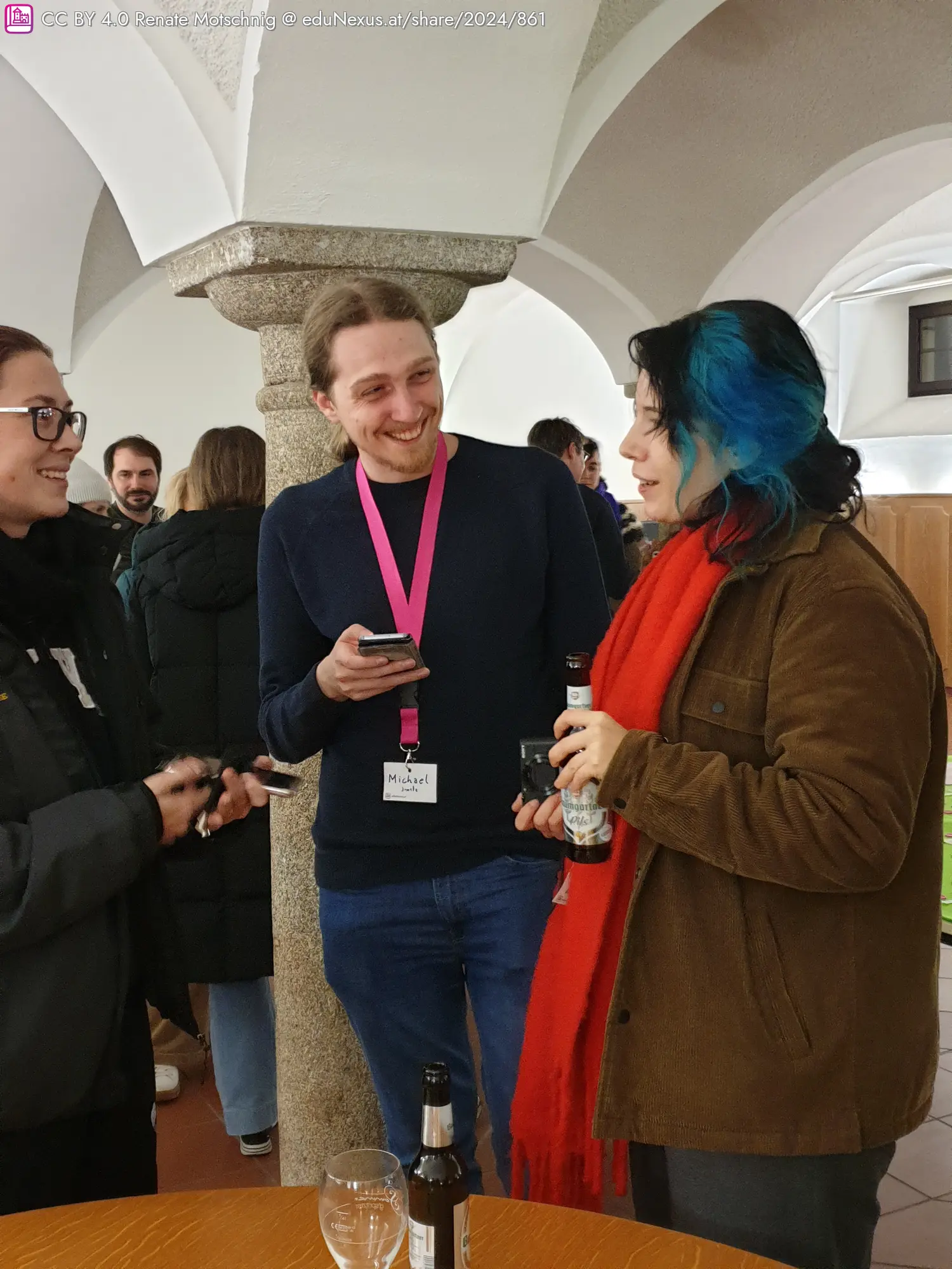 Drei Personen stehen an einem Tisch, zwei halten Smartphones, eine ein Getränk. Eine Person trägt ein pinkes Band mit Namensschild. Im Hintergrund sind weitere Personen und eine Säule sichtbar.