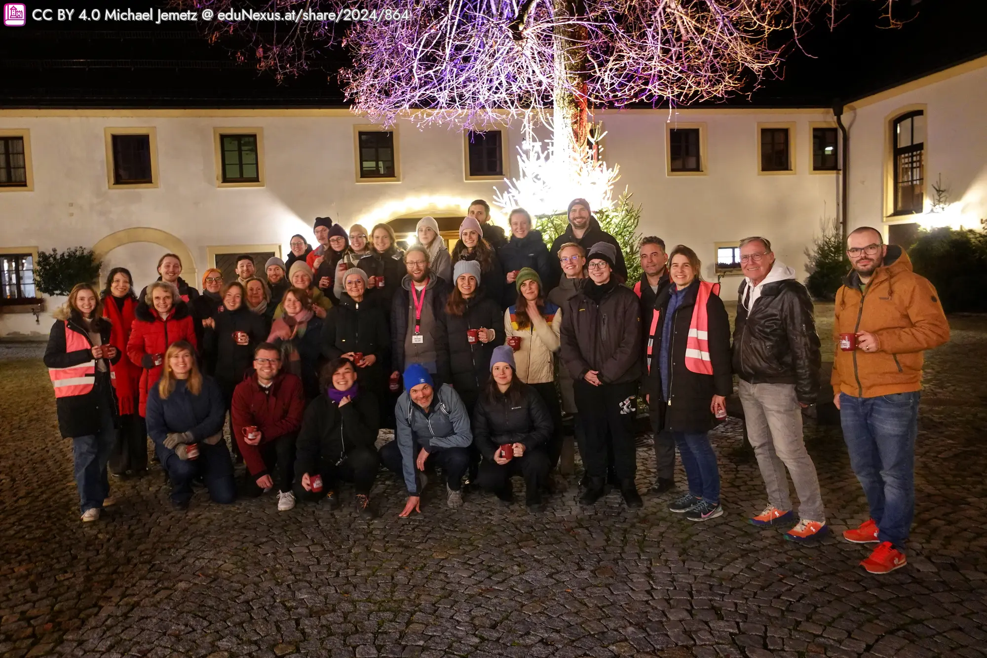 Gruppenfoto von etwa 40 Personen in einem Innenhof, beleuchtet von einem Weihnachtsbaum, während sie lächeln und zusammenstehen. Einige tragen rote Westen und Winterkleidung.